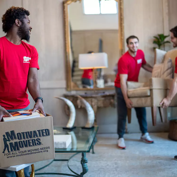 A few team members at Motivated Movers carefully lift a chair while another packs a customer's belongings into a moving box.